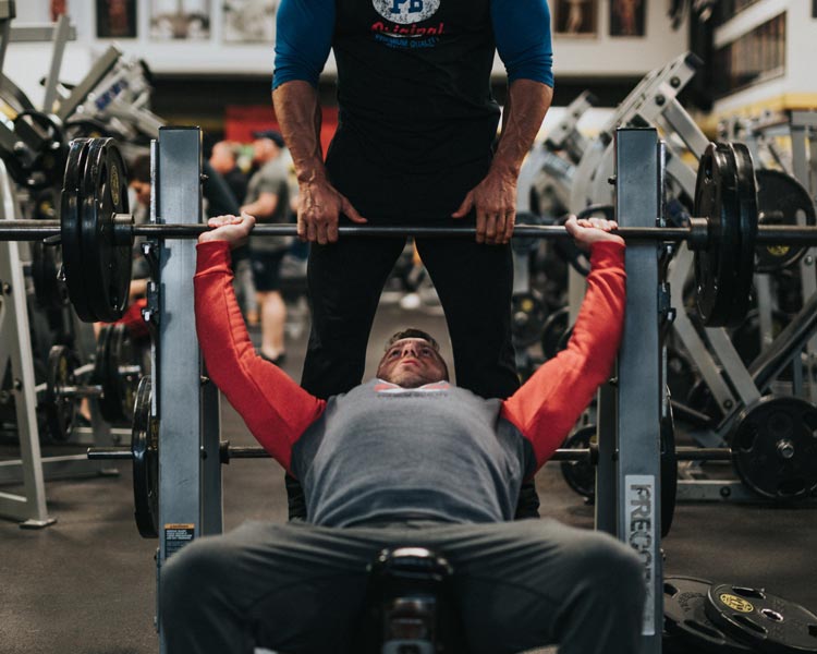 Online Fitness Plans -Man doing a bench press at the gym supported by personal trainer